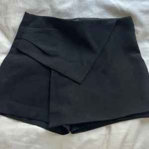 Svart skort från zara - Snygga svarta omlottshorts från Zara Basic med hög midja och stilren design. Shortsen har en omlott-detalj framtill, diskreta fickor bak och dragkedja i sidan. Har tyvär ett litet hål