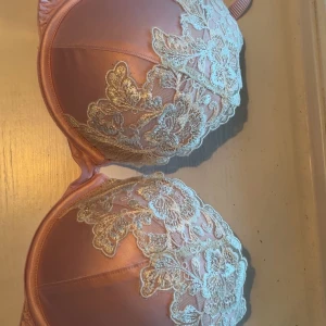 Rosa spets-bh från Ella M. - Supersöt rosa bh från Ella M. med vacker blommig spets i vitt över kuporna. Bh:n har justerbara axelband och en mjuk satinliknande yta som känns lyxig. Perfekt för dig som gillar romantiska detaljer och en feminin look. Mycket bra skick, använd enstaka gånger! Nypris 499kr