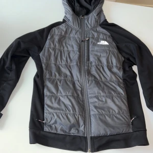 Svart och grå pufferjacka från The North Face - Snygg svart och grå pufferjacka från The North Face med huva och hel dragkedja. Jackan har quiltad framsida, mjuk fleece på insidan och två sidofickor med dragkedja. Perfekt för kyliga dagar och outdoor vibes.