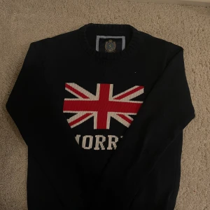 Mörkblå stickad tröja från Morris - Snygg mörkblå stickad tröja från Morris med Union Jack-flagga och logga framtill. Tröjan har rund halsringning och långa ärmar, perfekt för dig som gillar klassisk brittisk stil. Passar dig som vill sticka ut med en preppy vibe.