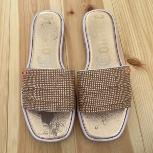 Glittriga sandaler från Juicy Couture - Snygga sandaler från Juicy Couture med glittriga rosa stenar över foten och fyrkantig tå. Ovandelen är dekorerad med små glittrande detaljer och har en metallbricka med logga. Sulan är ljusrosa och skon har platt sula, perfekt för sommardagar.