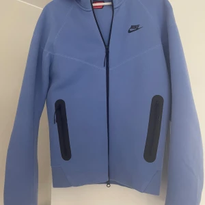 Blå Nike hoodie med dragkedja - Snygg blå hoodie från Nike med hel dragkedja framtill och svarta detaljer vid fickorna. Tröjan har huva, långa ärmar och en stilren look med Nike-logga på bröstet. Perfekt för en sportig och avslappnad stil.