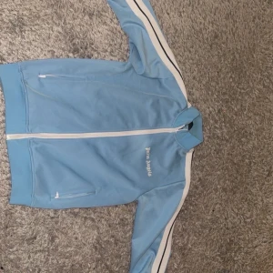 Blå track jacket från Palm Angels - Snygg ljusblå track jacket från Palm Angels med vita och svarta ränder längs ärmarna. Jackan har hel dragkedja framtill, ståkrage och två fickor med dragkedja. Perfekt för en sportig och trendig look.
