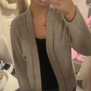 Beige stickad kofta med håldetaljer - Säljer en beige stickad kofta med snygga håldetaljer längs kanterna. Koftan har en öppen front och långa ärmar, perfekt att slänga på sig över en topp. Enkel och stilren design som passar till mycket.