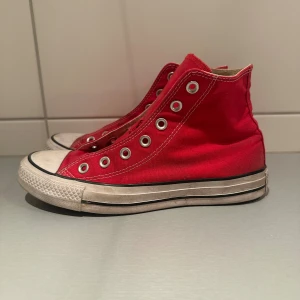 Röda Converse Chuck Taylor All Star - Klassiska röda Converse Chuck Taylor All Star high-tops med vit gummisula och rund tå. Skorna är i canvas med vita kontrastsömmar och metallöljetter för snörning. Perfekta för en avslappnad och cool streetstil.