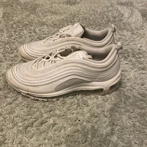 Nike Air Max 97 helvita sneakers - Säljer ett par Nike Air Max 97 i helvitt med ikoniska vågformade paneler och synlig Air-sula. Skorna har snörning, rund tå och är tillverkade i syntet och textil. Perfekta för dig som gillar en clean och sportig look.