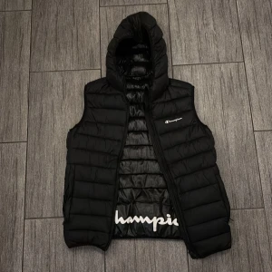 Svart dunväst från Champion med huva - Säljer en svart dunväst från Champion med huva och dragkedja. Västen är knappt använd och är i nytt skick. Säljer den billigt då den tar upp plats i garderoben. (Ny pris 700) Hör av dig om du är intresserad. Mvh Gustav