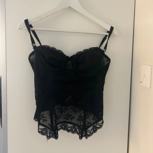 Svart spetskorsett med rosett - Säljer en svart korsett med justerbara axelband, vacker spets och liten rosett framtill. Toppen har genomskinliga spetsdetaljer med blommönster och hakknäppning bak. Perfekt för dig som vill ha en edgy och feminin look.