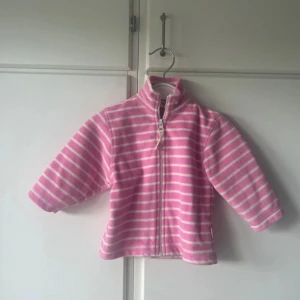 Rosa fleecejacka från Polarn O. Pyret - Mjuk och skön rosa fleecejacka från Polarn O. Pyret med vita horisontella ränder. Jackan har hög krage, hel dragkedja framtill och långa ärmar. 