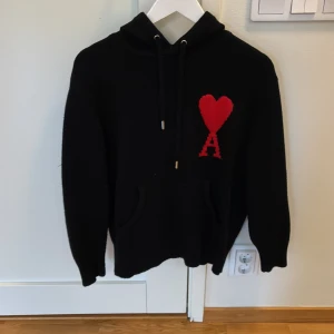 Ami Paris  Hoodie Svart Äkta Stickad - Svart hoodie från Ami med stor röd hjärtlogga och bokstaven A på bröstet. Tröjan har huva med snörning, känguruficka och är stickad i mjukt material. Perfekt för dig som gillar streetstyle och vill sticka ut med en ikonisk detalj.