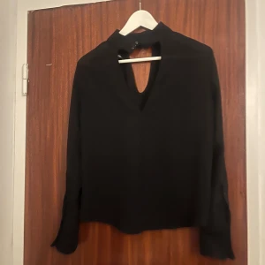 Svart blus med öppen rygg från H&M Divided - Svart blus från H&M Divided med lång ärm och snygg öppen rygg. Hög hals med tre dekorativa knappar i nacken. Toppens material känns mjukt och lätt, perfekt för att lyfta din outfit med en stilren touch. Nedersta bandet till knappen är trasig (se bild) men går fortfarande att knäppa