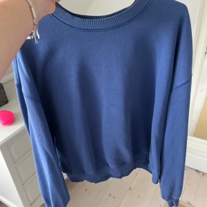 Blå sweatshirt med rund hals - En enkel och stilren blå sweatshirt med rundad hals och ribbade muddar vid ärmslut och nederkant. Tröjan har en loose passform och är långärmad, perfekt för chill dagar. Materialet känns mjukt och skönt mot huden.