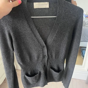 Mörkgrå kofta från Jan Machenhauer - Snygg mörkgrå kofta från Jan Machenhauer med v-ringning, två framfickor och knappar framtill. Koftan är långärmad och har en ribbad midja som ger en schysst passform. Perfekt för lager-på-lager och chill stil.