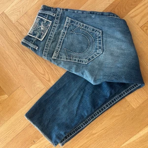 True Religion blå jeans  - Säljer ett par blå jeans från True Religion med tydliga kontrastsömmar och stora bakfickor med den klassiska hästsko-loggan. Passar 160-170cm. Skick 9/10. Äkta