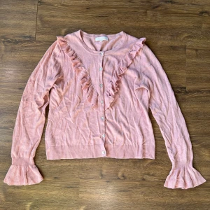 Rosa kofta med volang från RoseBud - Supersöt ljusrosa kofta från RoseBud med volangdetaljer längs framstycket och vid ärmsluten. Koftan har rund hals, knappar framtill och är stickad i mjukt material. Perfekt för dig som gillar romantisk och feminin stil.