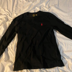 Svart långärmad tröja Polo Ralph Lauren - Svart långärmad tröja från Polo Ralph Lauren med klassisk röd logga broderad på bröstet. Tröjan är i mjuk bomull och har en rund halsringning. Perfekt för dig som gillar stilrena och tidlösa plagg med en sportig touch.