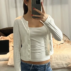 Vit stickad väst med knappar - Säljer en söt vit stickad väst med diskret hålmönster och rund hals. Västen är croppad och har knäppning framtill med små vita knappar. Perfekt att slänga över ett linne för en fräsch look. Enkel och stilren design som passar till jeans eller kjol.