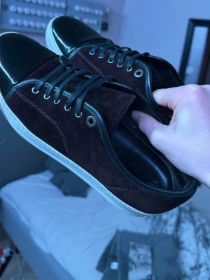 Vinröda sneakers från Lanvin med lackdetalj - Säljer ett par stilrena sneakers från Lanvin i vinröd mocka med svarta lackade tår och svarta skosnören. Skorna har vit sula och metallfärgade öljetter. Perfekta för dig som vill ha en lyxig touch på vardagslooken. Storlek 44. Skick 10/10 knappt använda. Hör av dig vid frågor!🙏🏻 OBS ingen sulkräm har använts!