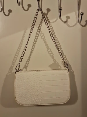 Vit axelväska med kedja - Snygg vit axelväska i croco-mönstrad syntet med silverfärgad kedjerem. Väskan har en stilren, rektangulär form och stängs med dragkedja upptill. Perfekt accessoar för att lyfta din outfit.