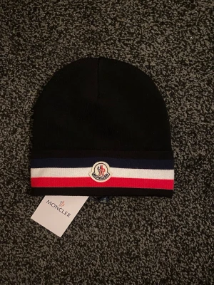 Moncler mössa - Snygg svart Moncler-mössa med klassiska ränder i blått, vitt och rött längst ner. Mössan har Moncler-logga framtill och är tillverkad i mjukt material som sitter skönt på huvudet. Perfekt accessoar för att lyfta din vinterstil.