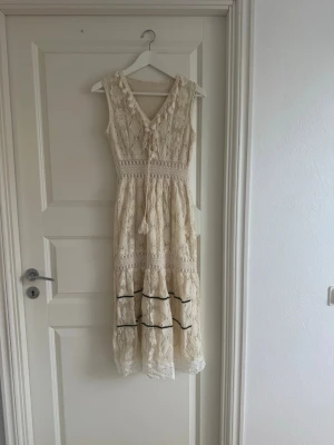 Spetsklänning med snörning - Romantisk långklänning i spets med blommigt mönster och virkade detaljer. Klänningen har v-ringning med snörning framtill, är ärmlös och har svarta dekorband nertill. Perfekt för sommarens alla tillfällen!