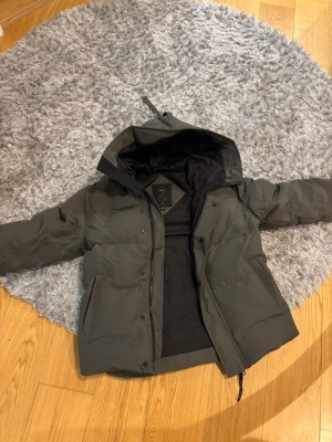Mörkgrå dunjacka från Canada Goose - Säljer en mörkgrå dunjacka från Canada Goose i storlek L. Jackan har huva, diskret logga på ärmen och dragkedja framtill. Fodrad för extra värme och har två stora fickor. Perfekt för kalla dagar och riktigt snygg streetstil. Kan tänkas gå ner i pris vid snabb affär 