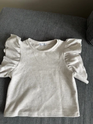 Beige topp med volangärmar från Zara - Supersöt beige topp från Zara med volangdetaljer på ärmarna. Toppen har rund halsringning och är gjord i mjuk bomull. Perfekt för dig som gillar en enkel men ändå trendig look. Användes ett fåtal gånger.