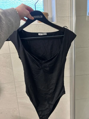 Svart body från Zara med rynk - Säljer en svart body från Zara med snygg rynkdetalj vid bysten och bredare axelband. Modellen har en djupare urringning och är tillverkad i ett mjukt, stretchigt material som sitter tight mot kroppen. Perfekt att styla med jeans eller kjol.