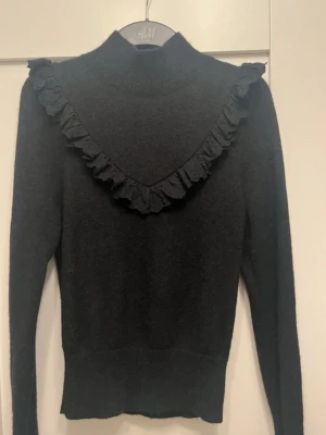 Svart polotröja med volang från & other stories - Säljer en svart polotröja från & other stories med lång ärm och snygg volangdetalj i V-form över bröstet och axlarna. 