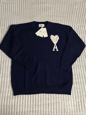 Ami Paris Tröja - Snygg mörkblå stickad tröja från Ami Alexandre Mattiussi med vit hjärtlogga och bokstaven A på bröstet. Tröjan har rund hals, ribbade muddar och är långärmad. Perfekt för dig som gillar stilrena och trendiga plagg. Tröjan är mjuk och kön och passar perfekt inför nuvarande vinter fast även kommande vår och sommar. Ett till ett. Tröja säljs på grund av fel storlek. Vid några funderingar är det bara att skriva. Skickar samma dag. 