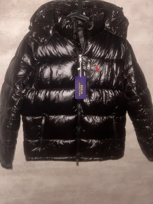 Svart pufferjacka från Polo Ralph Lauren - Säljer en svart, glansig pufferjacka från Polo Ralph Lauren med röd logga på bröstet. Jackan har huva, flera dragkedjor och quiltad design. Perfekt för kalla dagar och riktigt snygg till streetwear-looken.