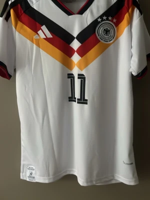 Tysklands fotbollströja Adidas L - Säljer en officiell Tyskland fotbollströja från Adidas i storlek L. Tröjan är vit med svarta, röda och gula detaljer, nummer 11 och namnet Woltemade på ryggen. Klassisk V-ringning och broderad landslagsemblem på bröstet. Perfekt för dig som älskar fotboll och snygg design.