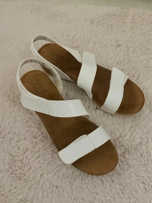 Vita sandaler - Snygga vita sandaler med breda remmar och kardborreknäppning. Sulan är i kork-look och innersulan är mjukt brun. Perfekta för sommaren! Fint skick!