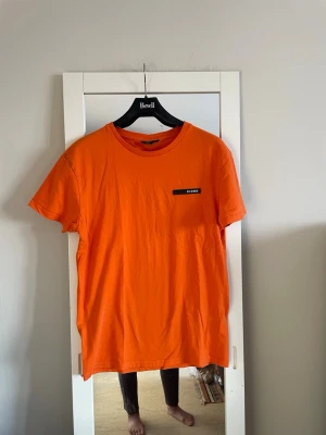 Orange t-shirt från ICONO, slim fit - Snygg orange t-shirt från ICONO i slim fit-modell. Klassisk rund hals och korta ärmar. T-shirten har en svart patch med vit ICONO-logga på bröstet. Tillverkad i mjuk bomull som känns skön mot huden.