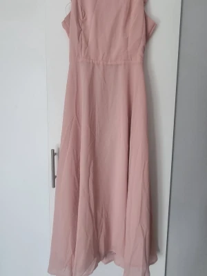 Ljusrosa långklänning med rosett - Superfin ljusrosa långklänning med tunna axelband och öppen rygg med en snygg rosettdetalj. Klänningen har ett flowy fall och är gjord i ett lätt, tunt material som känns luftigt. Perfekt för sommarens alla tillfällen!