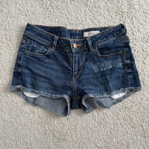 Mörkblå jeansshorts &SHORTS strl 34 - Säljer ett par mörkblå jeansshorts från &SHORTS i storlek 34. De har råa, fransiga kanter, klassisk femficksdesign och en liten patchdetalj fram. Perfekta för sommaren och riktigt snygga till sneakers eller sandaler.
