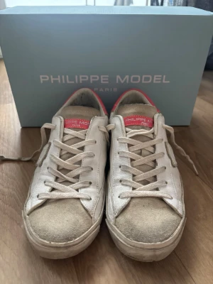 Vita Philippe Model sneakers med rosa detalj - Säljer ett par vita Philippe Model sneakers i skinn med beige mockadetaljer och rosa hälparti. Skorna har snörning, rund tå och platt sula. Klassisk logga på sidan och bak på hälen. För dom som inte vet ska modellen på skorna vara ”smutsiga”. Har använt dom en sommar, säljer då dom är lite förstora för mig. Köpte för 3500kr