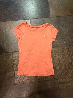 Korallfärgad t-shirt från Polo Ralph Lauren - Snygg korallfärgad t-shirt från Polo Ralph Lauren med klassisk gul broderad logga på bröstet. T-shirten har rund halsringning och korta ärmar. Perfekt till jeans eller shorts för en clean och enkel look.