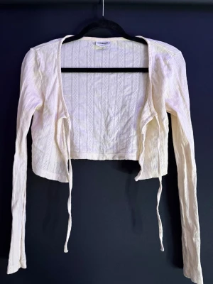 Vit croppad kofta från Noisy May - Säljer en vit croppad kofta från Noisy May med långa ärmar och snyggt ribbat mönster. Koftan har knytband framtill och är tillverkad i ett mjukt, tunt material som passar perfekt till lager på lager. Enkel att styla till olika outfits!