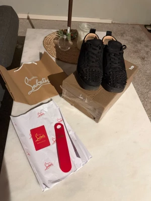 Svarta Christian Louboutin sneakers med nitar - Säljer ett par svarta sneakers från Christian Louboutin med ikoniska röda sulor och coola svarta nitar över tån. Skorna har snörning och är tillverkade i mocka för en lyxig känsla. Perfekta för dig som vill sticka ut med en edgy och exklusiv look. Några nitar saknas då de är använda mycket men annars Perrfa 