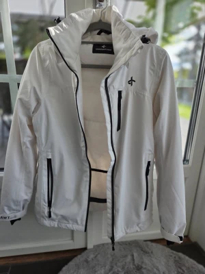 Vit vindjacka/skidjacka från Cross Sportswear - Vit vindjacka/skidjacka från Cross Sportswear med svarta detaljer och dragkedjor. Jackan har huva, flera praktiska fickor med dragkedja och justerbara ärmslut. Tillverkad i 100% polyester. 