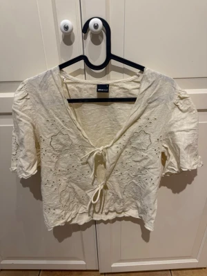 Ljusgul blus med broderi från Gina Tricot - Säljer en kortärmad blus från Gina Tricot med broderi. Blusen har korta puffärmar, knyt framtill och volangkant vid ärmslut och nederkant. 