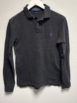 Mörkgrå långärmad henley från Polo Ralph Lauren - Snygg mörkgrå långärmad henley från Polo Ralph Lauren i slim fit. Skjortan har klassisk krage, två knappar vid halsen och en lila broderad logga på bröstet. Tillverkad i mjuk bomull som känns skön mot huden. Perfekt för dig som gillar stilrena plagg med en twist. 100% äkta. Skriv för frågor!