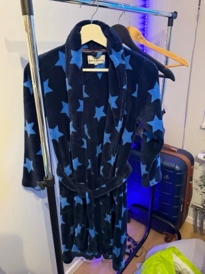 Mysig blå fleeceväst med stjärnor - Säljer en fluffig och skön blå fleeceväst från Hampton Republic med stora ljusblå stjärnor över hela västen. Västen har skärp i midjan och bred krage för extra mysfaktor. Perfekt för chill dagar hemma eller när du vill ha något riktigt bekvämt.