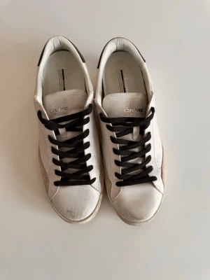 Vita sneakers från Crime London - Snygga vita sneakers från Crime London med svarta skosnören och detaljer. Skorna har en klassisk låg profil, rund tå och platt sula i gummi med brunt mönster undertill. Ovandelen är i skinn och de har stilrena sömmar på sidorna.Ser mer används ut än vad dom är för att dem är distressed, används endast ett par gånger 