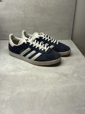 Adidas Gazelle blåvita sneakers - Säljer ett par klassiska Adidas Gazelle sneakers i mörkblå mocka med vita detaljer och de ikoniska tre ränderna på sidan. Skorna har vit sula, snörning och guldfärgad Gazelle-text på sidan. Perfekta för dig som gillar retrostil och streetwear.