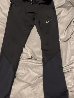 Svarta träningsbyxor Nike XL - Säljer ett par svarta träningsbyxor från Nike i storlek XL. Byxorna har dragkedja vid benslut, liten ficka med dragkedja bak och justerbar midja med snörning. Tillverkade i ett stretchigt syntetmaterial med diskret Nike-logga på låret.