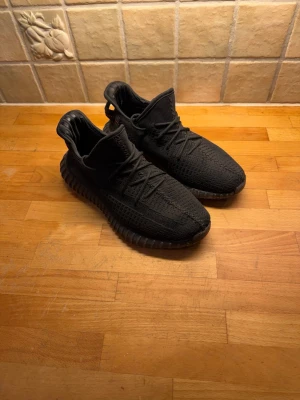 Svarta Adidas Yeezy Boost 350 V2 - Säljer ett par svarta Adidas Yeezy Boost 350 V2 med stickad ovandel och klassisk ribbad sula. Skorna har snörning, rund tå och en diskret röd detalj på hälen. Superbekväma och ikoniska sneakers som passar perfekt till streetwear.