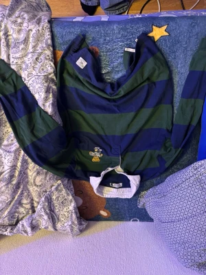 Randig rugbytröja Beverly Hills Polo Club - Säljer en klassisk rugbytröja från Beverly Hills Polo Club i storlek M. Tröjan har breda marinblå och gröna ränder, vit krage och en broderad nalle med rugbyboll på bröstet. Tillverkad i mjuk bomull och har långa ärmar. Perfekt för en sportig och avslappnad stil.