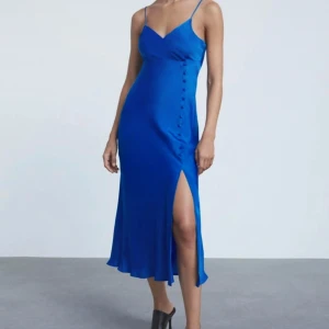 Blå sidenklänning med slits - Snygg blå långklänning i siden med smala axelband och v-ringning. Klänningen har en slits på sidan och dekorativa knappar längs höften. 